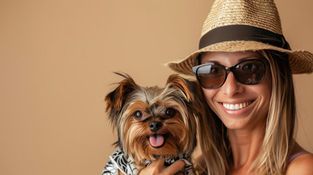 Woman smiling with her Yorkshire Terrier puppy dressed in a funny outfit, including a hat and sunglasses, against a beige studio backdrop. Space for text on the left.の素材