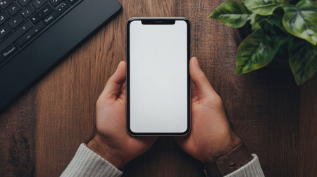 Man hand with a blank white screen phone, perfect for advertisement, on a wooden home office desk.の素材