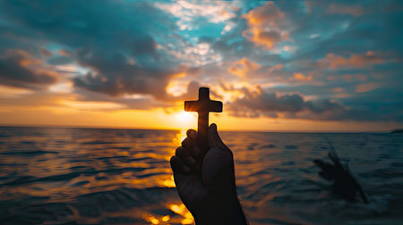 Silhouette of a cross held in a human hand against a vibrant sunrise, symbolizing faith and hope with this inviting photo.の素材