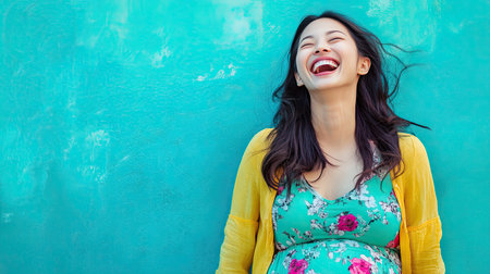 A joyful portrait of a pregnant Asian woman laughing, with a bright blue background providing a cheerful setting, perfect for maternity and family-related themes.の素材