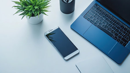 Modern office scene with a blue MacBook, smartphone, and plant on a white desk. Clean lines and a pop of color create a fresh, tech-savvy environment.の素材
