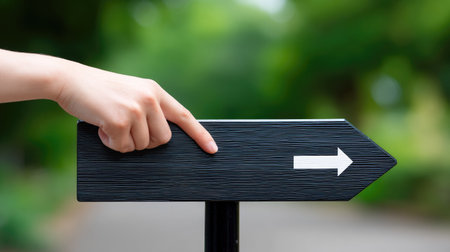 A close-up of a hand pointing towards a directional arrow sign on a pole, set against a blurred green nature background, symbolizes guidance and decision-making.の素材