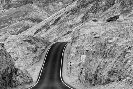 Road through Artists Drive in Death Valley National Park that leads over a hill and seems to disappear in the mountains (black and white photo)の写真素材