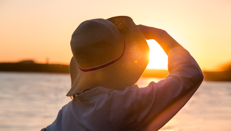 Young and beautiful woman wearing a hat in sunset light looking far away. Photo from behindの写真素材
