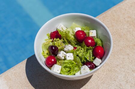 Fresh and healthy salad in a isolated white bowl. At the side of the swimming pool, in a sunny day. Lettuce, seeds, big red and juicy cherries, feta cheese and olive cold pressed oil.の写真素材