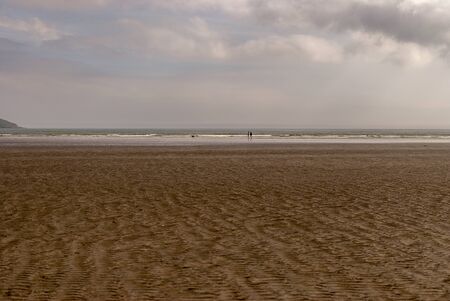 Couple with a dog walking and playing on sandy beach leaving footprints in the sand. Family enjoying the day. Cloudy day.の写真素材