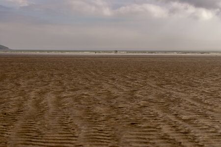 Couple with a dog walking and playing on sandy beach leaving footprints in the sand. Family enjoying the day. Cloudy day.の写真素材