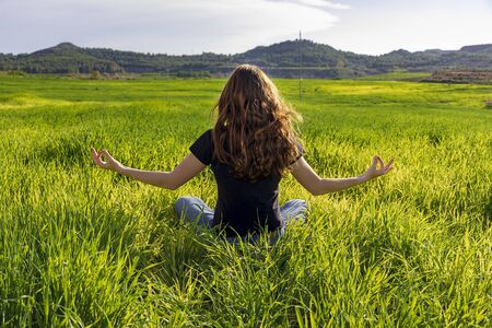 Young caucasian woman red-haired with freckles, resting on a green field at spring sunset, sitting in a yoga position. Meditation, mindfulness, relaxの写真素材