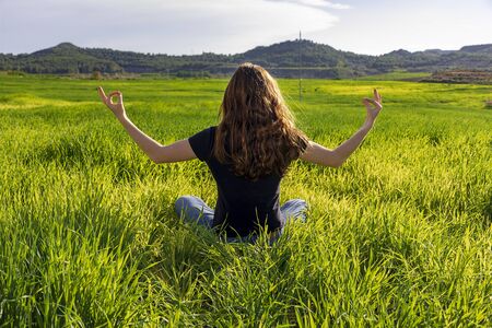 Young caucasian woman red-haired with freckles, resting on a green field at spring sunset, sitting in a yoga position. Meditation, mindfulness, relaxの写真素材