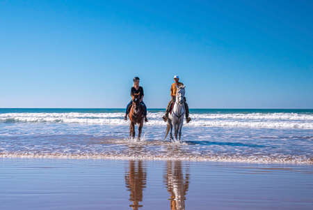 Man and woman riding horses along shoreline at beach against clear sky