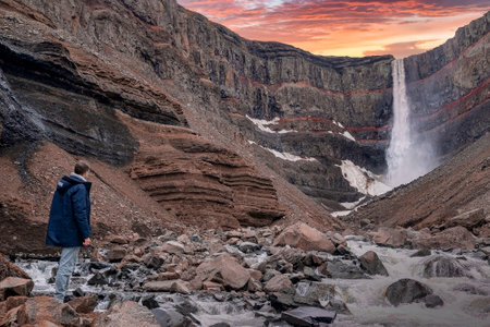 Male tourist exploring beautiful Hengifoss waterfall against sky during sunsetの写真素材