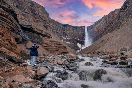 Male tourist with arms outstretched looking at Hengifoss waterfall during sunsetの写真素材