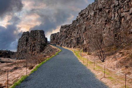 People walking on road by cliffs against cloudy sky at Thingvellir national parkの写真素材