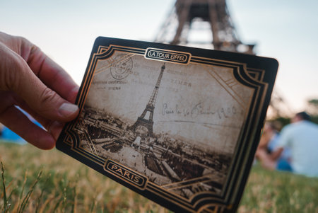 A hand holding an old vintage postcard featuring the Eiffel Tower, with the actual tower visible in the sunny background.の写真素材