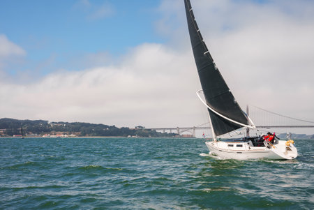 Sailing yacht with black sail near SF Oakland Bridge in SF Bay.
