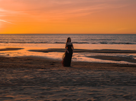Happy beautiful woman in a long black dress enjoying beautiful sunset on the Beach in California.の写真素材