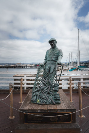 Life Size Fisherman Statue on Wooden Pier, Coastal Setting with Sailboatsの写真素材