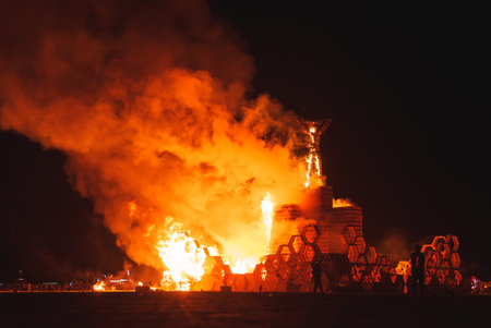 Dramatic night scene with burning wooden structure at desert art festival. Symbolizes impermanence, creativity, and community. Lights and people in background.の写真素材