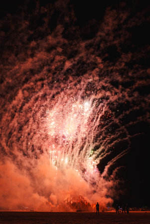 Vibrant fireworks illuminate night sky at music festival in desert setting, bursting pink, white, and green hues. Silhouettes of attendees marvel below.の写真素材