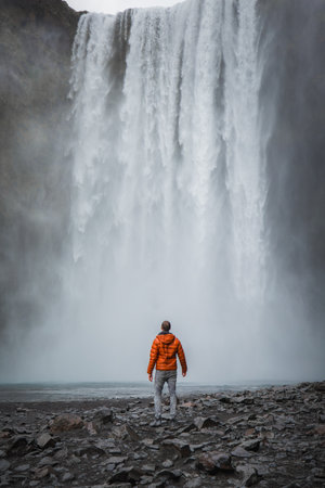 Person in Orange Jacket Facing Majestic Skogafoss Waterfall in Iceland