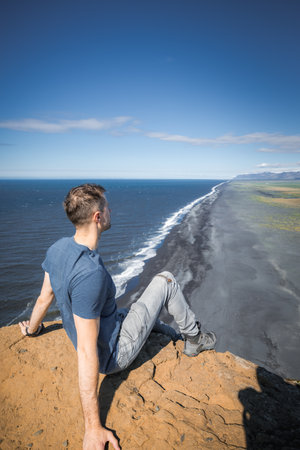 Man Sitting on Cliff Overlooking Reynisfjara Black Sand Beach in Iceland