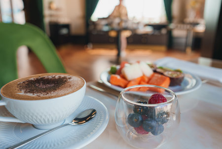A beautifully presented cappuccino in a white cup, accompanied by fresh berries and assorted fruits,...