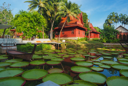 Traditional Thai Red Wooden Buildings with Pond and Palm Treesの写真素材
