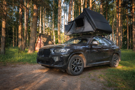 A black SUV with an open rooftop tent is parked in a forest. Tall trees surround the vehicle, with...