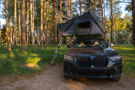A modern SUV with a rooftop tent and ladder is parked in a forest clearing. Tall trees with green foliage surround the serene camping setup.の写真素材