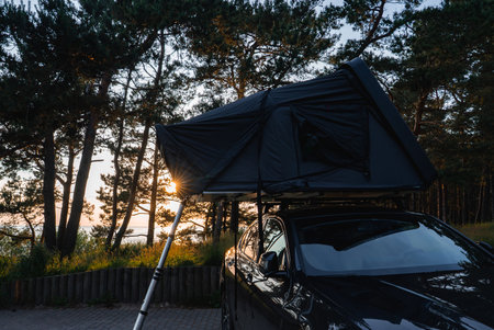 A modern SUV with a dark rooftop tent and ladder is parked in a forest. Tall trees surround the vehicle, with sunlight glowing through the canopy. A body of water is visible in the distance.の写真素材