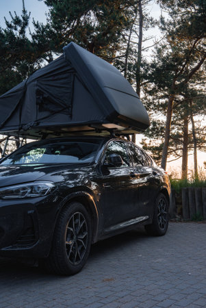 A modern black SUV with a partially open rooftop tent is parked on a paved surface in a forest, surrounded by tall trees under soft natural light.の写真素材