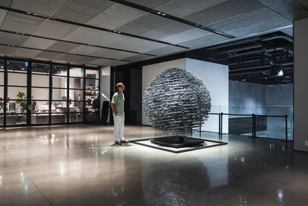 Spherical metallic sculpture in minimalist museum gallery, visitor observes