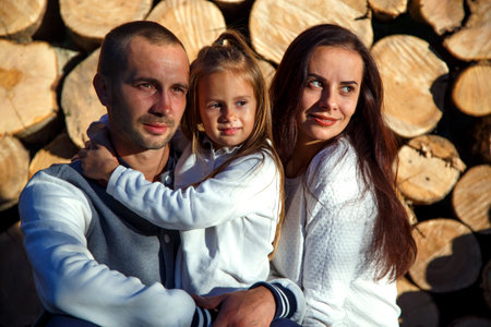 Young happy family on the background of firewood. Dad, mom and daughter are sitting near the chopped firewood on the farm. The concept of family, happiness, comfort and warmth.の写真素材
