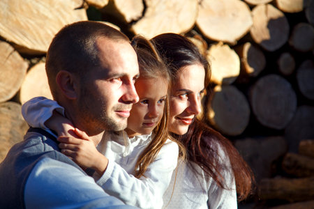 Young happy family on the background of firewood. Dad, mom and daughter are sitting near the chopped firewood on the farm. The concept of family, happiness, comfort and warmth.の写真素材