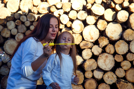 Mom and daughter blow bubbles on the background of firewood. Happy woman with child blow bubbles near stacked firewood. The concept of happiness, family, joy.の写真素材
