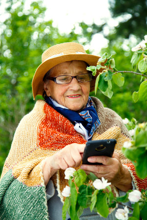Portrait of a grandmother in the garden taking a photo of herself on her phone. An old woman takes a selfie on her phone against the backdrop of flowering bushes.の写真素材
