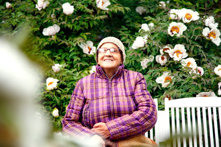 An old woman in a park on a bench with a tree peony flower. The concept of care, old age and happiness.の写真素材