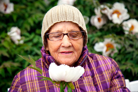 An old woman in a park on a bench with a tree peony flower. An old lady enjoys life. The concept of care, old age and happiness.の写真素材