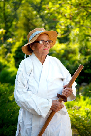 An old lady in a white robe in a park with a wooden stick. Portrait of an old lady posing with a wooden stick in the garden. The concept of old age, nature.の写真素材