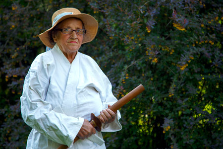 An old lady in a white gi in a park with a wooden sword. Portrait of an old lady training with a wooden sword in the garden. The concept of old age, sports, nature.の写真素材