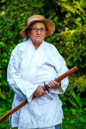 An old lady in a white robe in a park with a wooden sword. Portrait of an old lady training with a wooden sword in the garden. The concept of old age, sports, nature.の写真素材