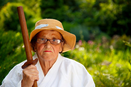 An old lady in a kimono in a park with a wooden sword. Portrait of an old lady training with a wooden sword in the garden. The concept of old age, sports, nature.の写真素材
