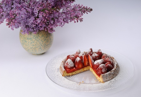Strawberry torte with fresh strawberries and powdered sugar on a glass plate next to a bouquet of lilacsの写真素材