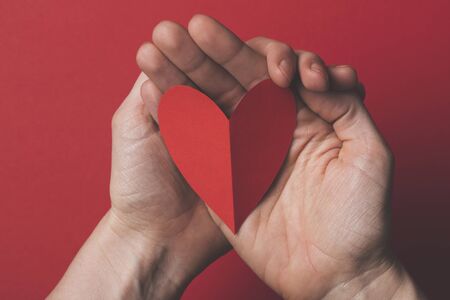 Female hand holding a red paper cut out heart on a plain red background.の写真素材