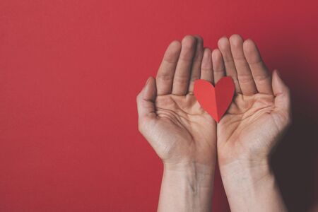 Female hand holding a red paper cut out heart on a plain red background.の写真素材