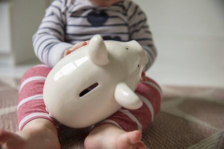 Cost of childcare concept. Young baby holds onto a white piggy bankの写真素材