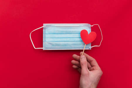 Hand holding a red love heart over a covid protective face mask.