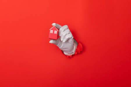 Santa Claus hand holding model house through a hole in red paper background
