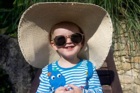 Cute little toddler wearing an oversized sun hat and sunglasses relaxing in the sunの写真素材