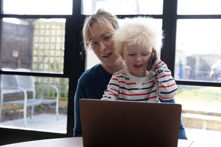 A mother working from home at a laptop computer with her young son on her lapの写真素材